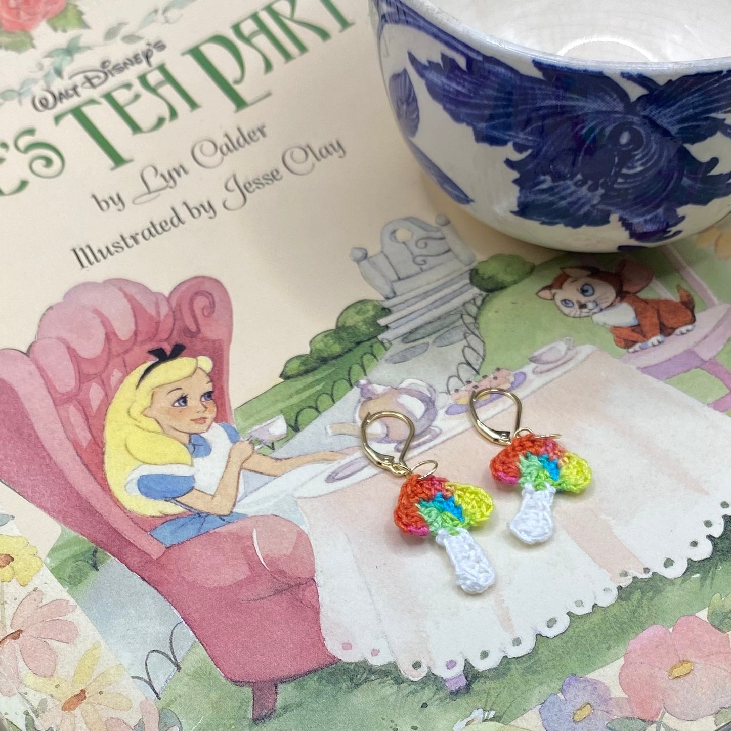 Rainbow-themed earrings on a Disney Alice in Wonderland book with a teacup in the background.
