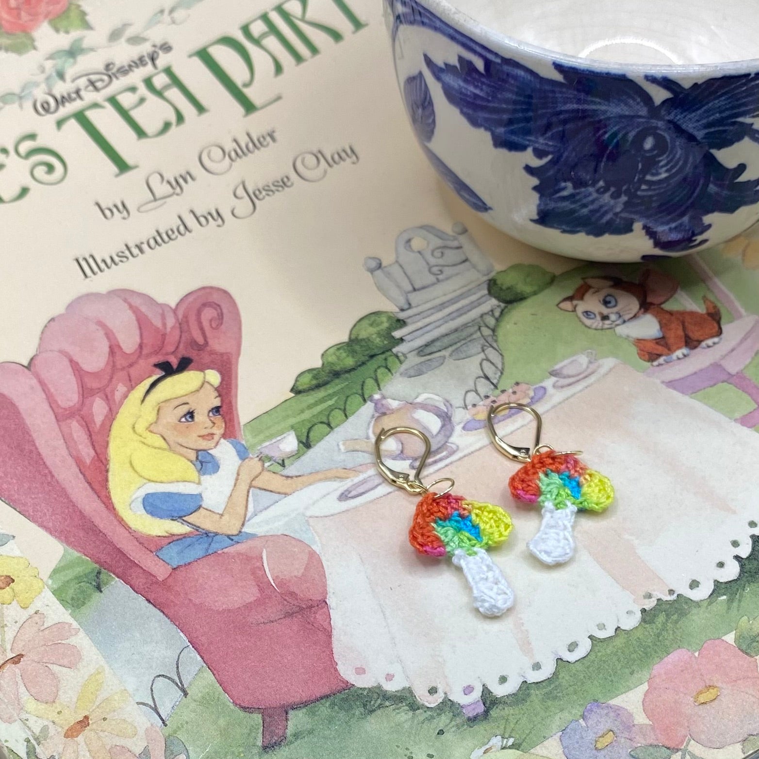 Rainbow-themed earrings on a Disney Alice in Wonderland book with a teacup in the background.