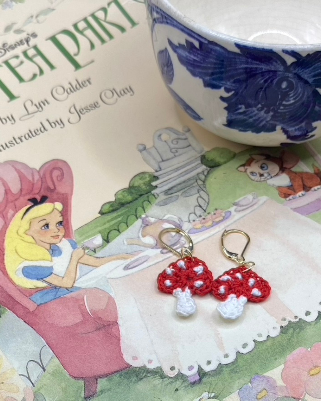 Red and white mushroom earrings on a Disney Alice in Wonderland tea party book with a teacup.