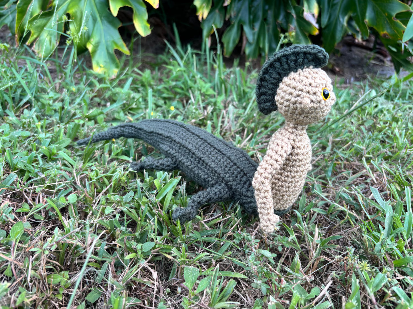 Crocheted alligator and person-like figure on grass with plants in the background