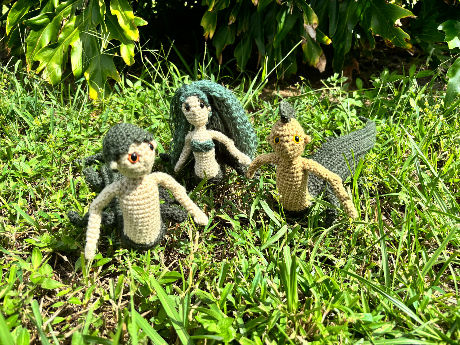 Three crochet mergators on grass with greenery in the background